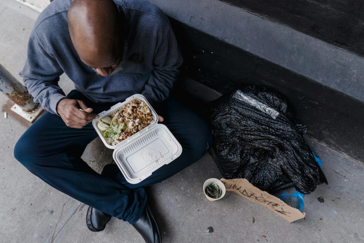 A man eats from a takeaway container while sitting on the sidewalk, highlighting urban hardship.