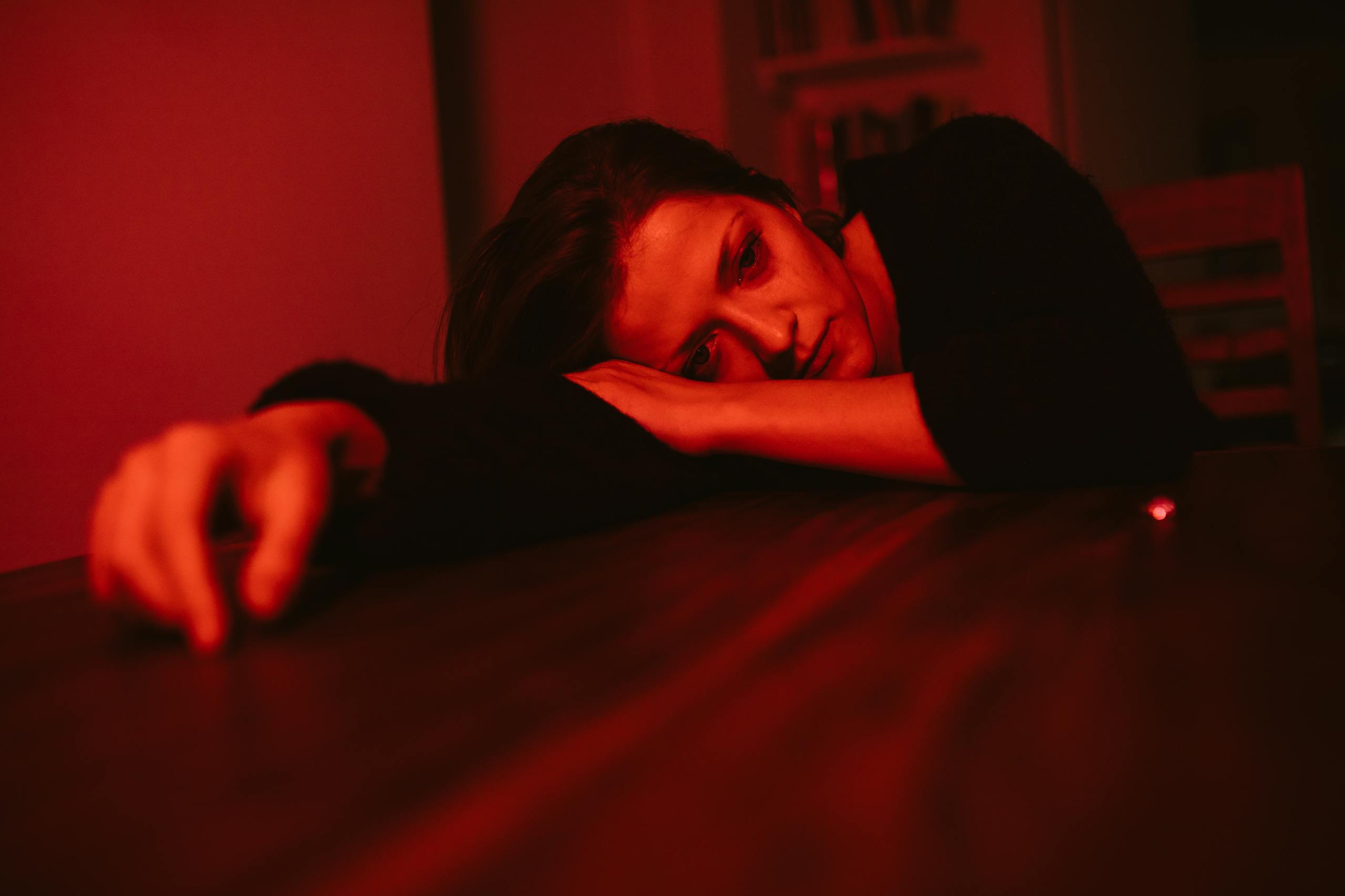 A pensive woman rests her head on a table, bathed in red lighting, reflecting emotion.