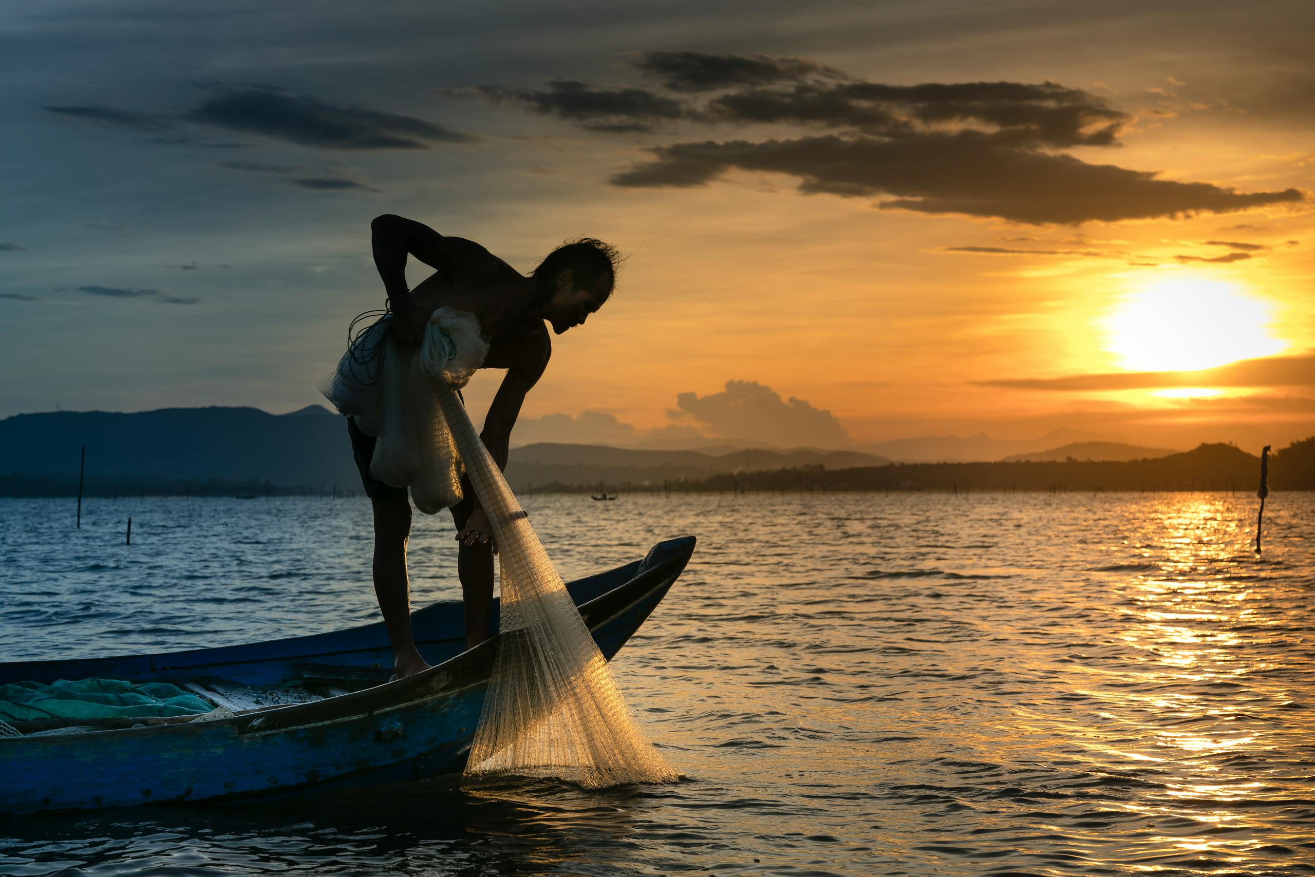 A silhouette of a fisherman on a boat casting a net during a serene sunrise over the sea.