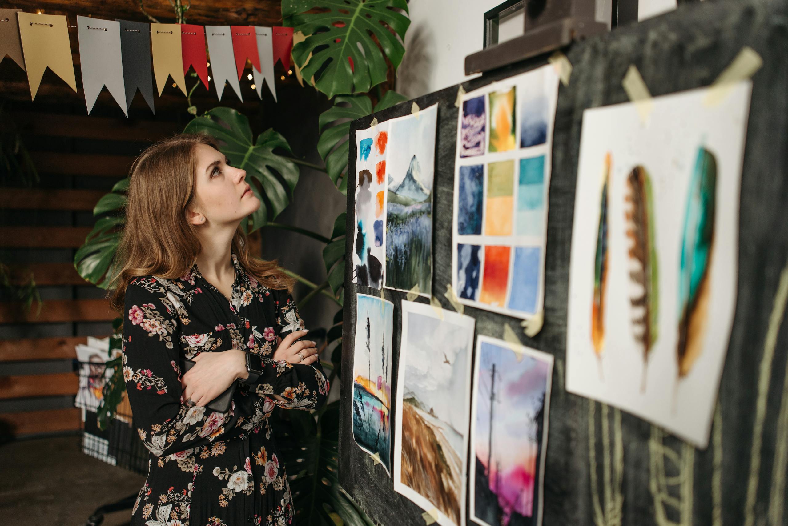 A woman in a floral dress observes art pieces on a board in her creative workshop.