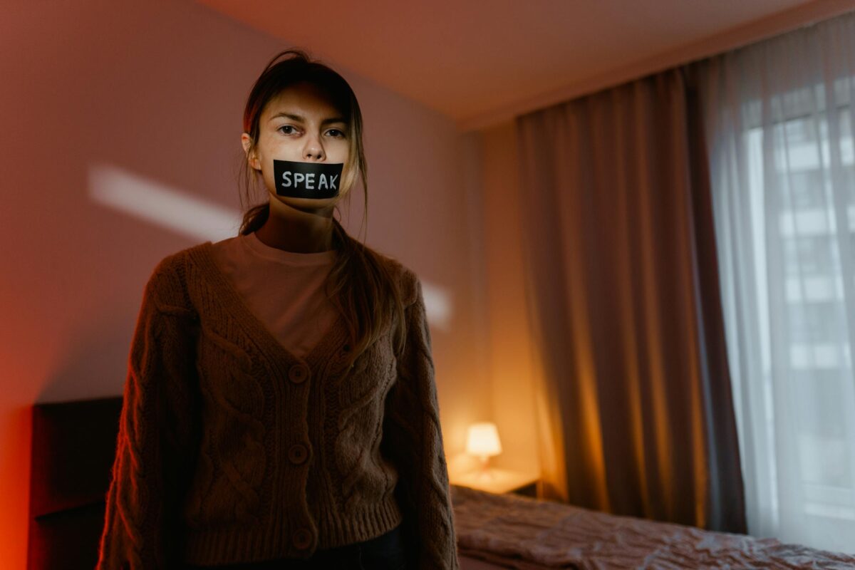 A woman stands in a dimly lit bedroom with tape reading 'Speak' over her mouth, symbolizing silence and oppression.
