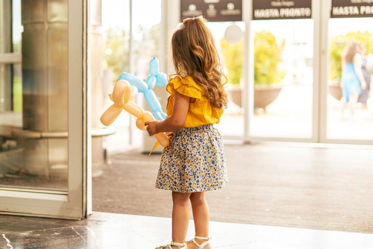 A young girl holding colorful balloon animals stands near automatic doors in a shopping mall.