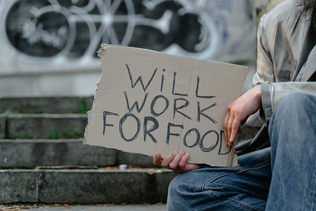 Pensive scene of a homeless person holding a 'Will Work for Food' sign on urban steps.