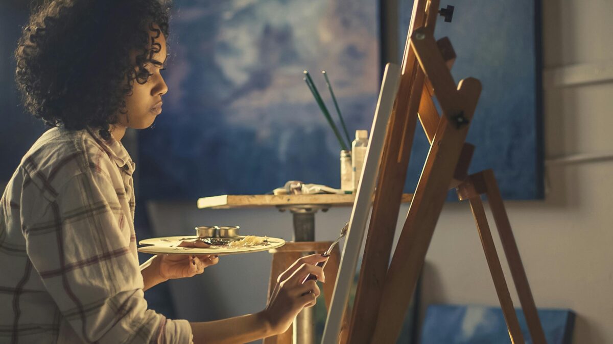 Side view of a young woman artist painting on canvas in a cozy studio setting.